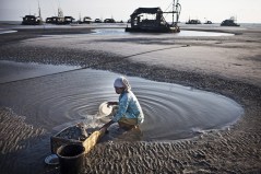 Een vrouw zoekt op Banka tin met de hand, terwijl op de achtergrond de machines van de grote bedrijven staan.